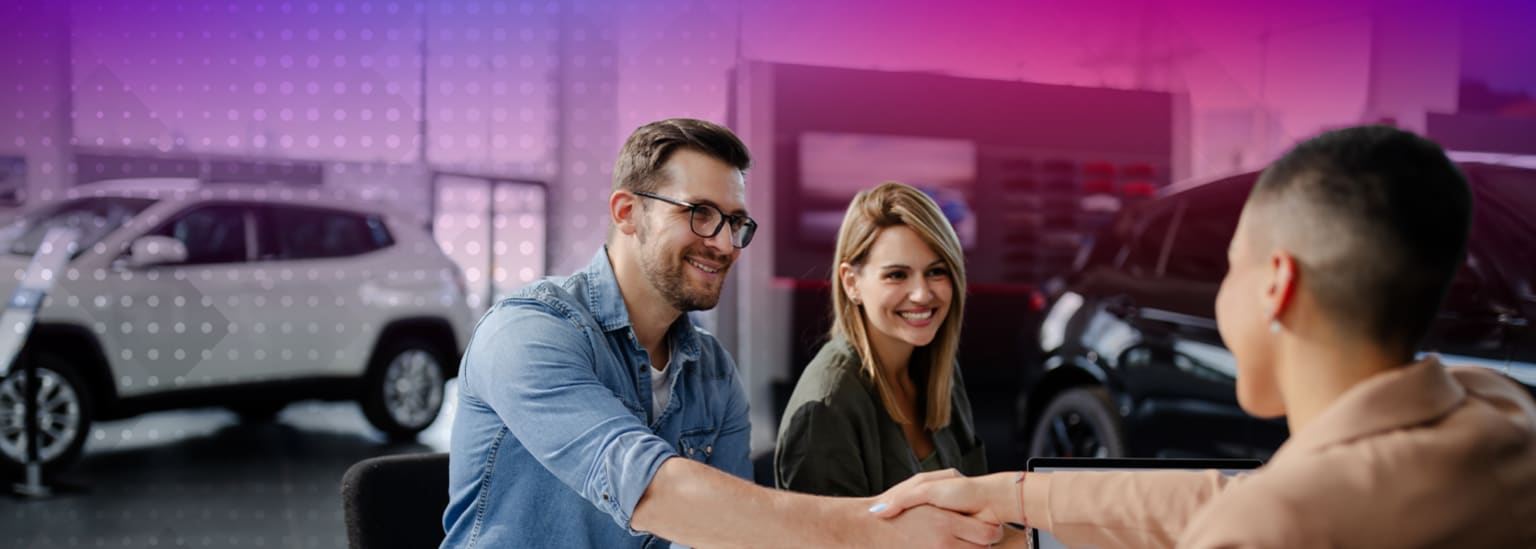 A smiling man and woman shaking hands in front of a colorful, illuminated backdrop with vehicles in the background.