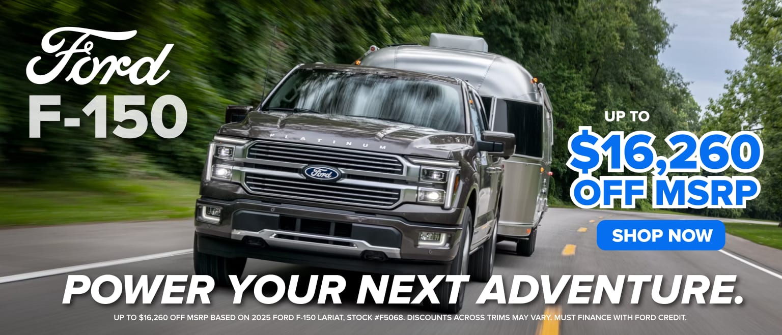 The image depicts a large, silver pickup truck driving on a road surrounded by lush, green trees. The truck is prominently featured in the foreground, with a promotional advertisement for the vehicle displayed on its side.