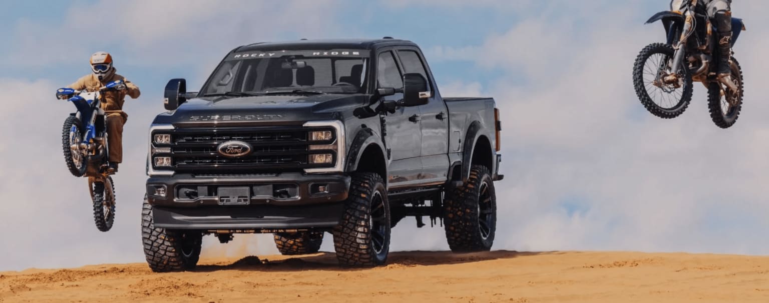 A large, heavily modified pickup truck with massive off-road tires is driving through a sandy desert landscape against a backdrop of a cloudy sky.