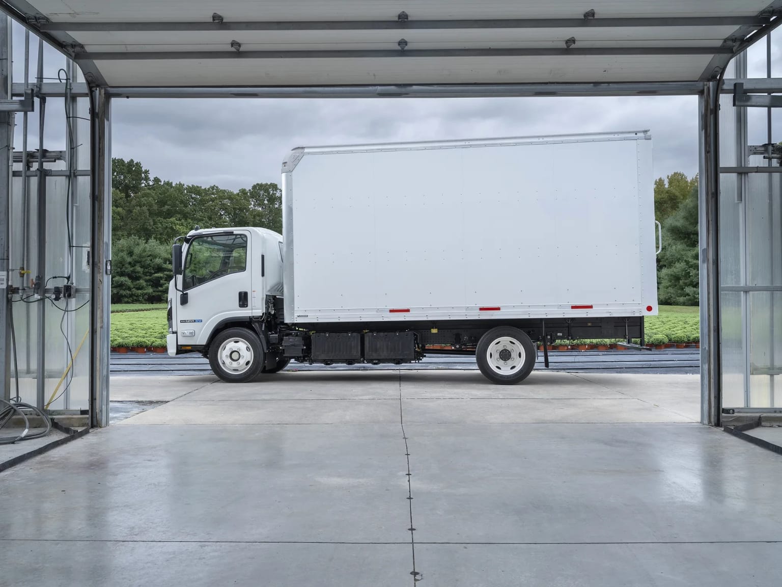 A white box truck parked inside a garage with a wet floor, trees and a cloudy sky in the background.