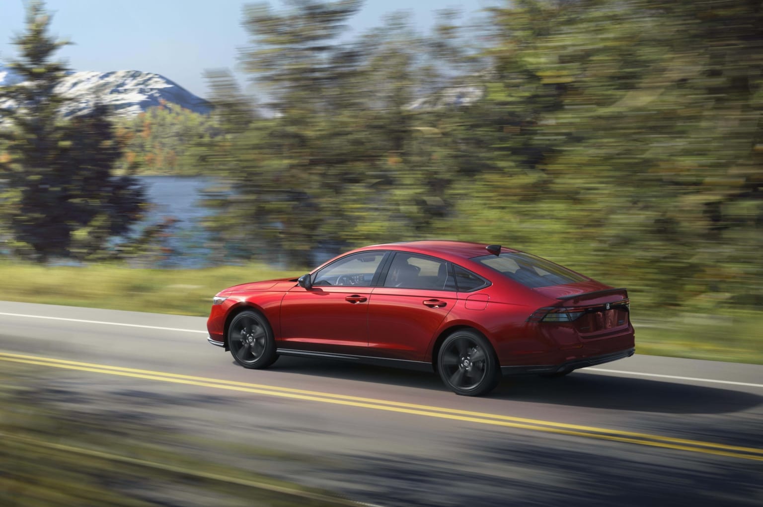 A red sedan driving on a winding road surrounded by lush green trees with a snow-capped mountain in the distance.