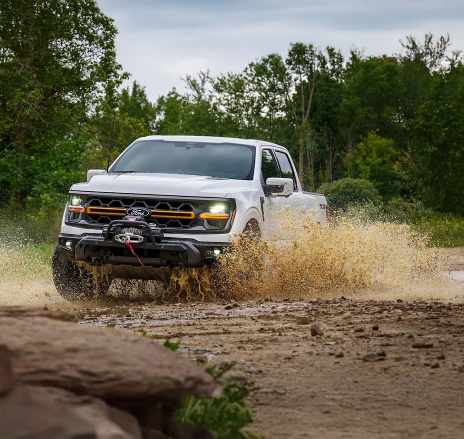 Off-road pickup truck driving through a muddy dirt road surrounded by green trees.