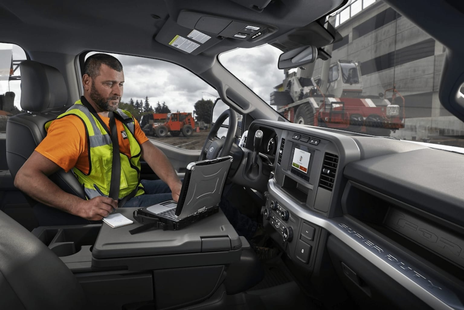 Interior of vehicle with person wearing high-visibility vest operating device, construction work in background with various equipment and vehicles visible.