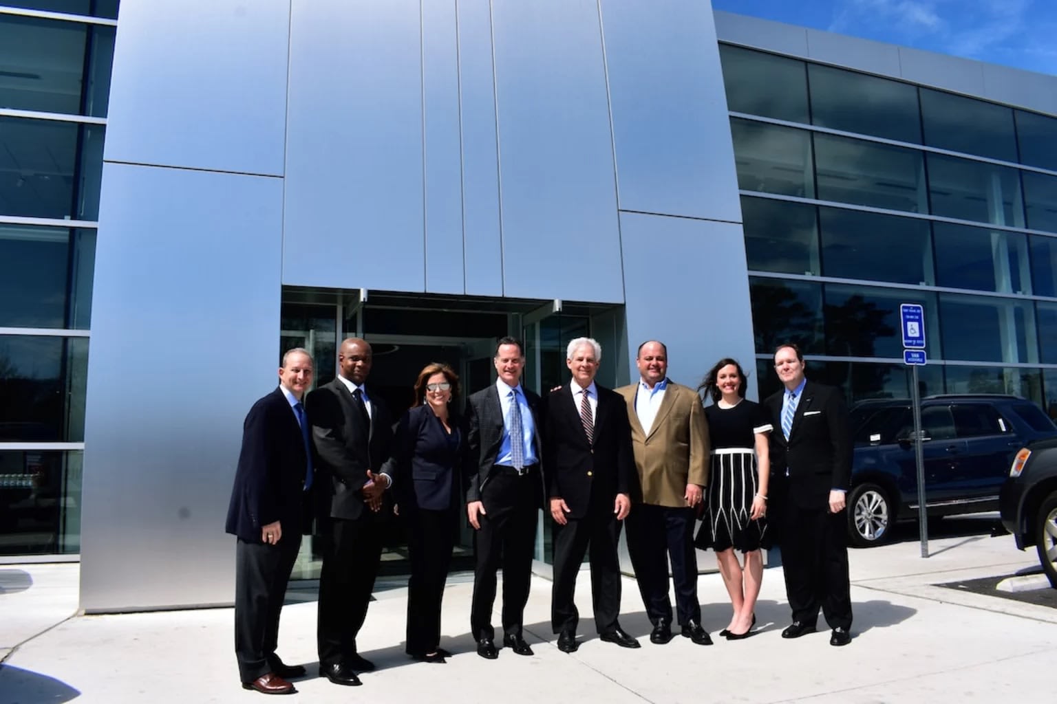 A group of people in formal attire stand in front of a modern silver building with large glass windows, while a dark SUV is parked to the right.