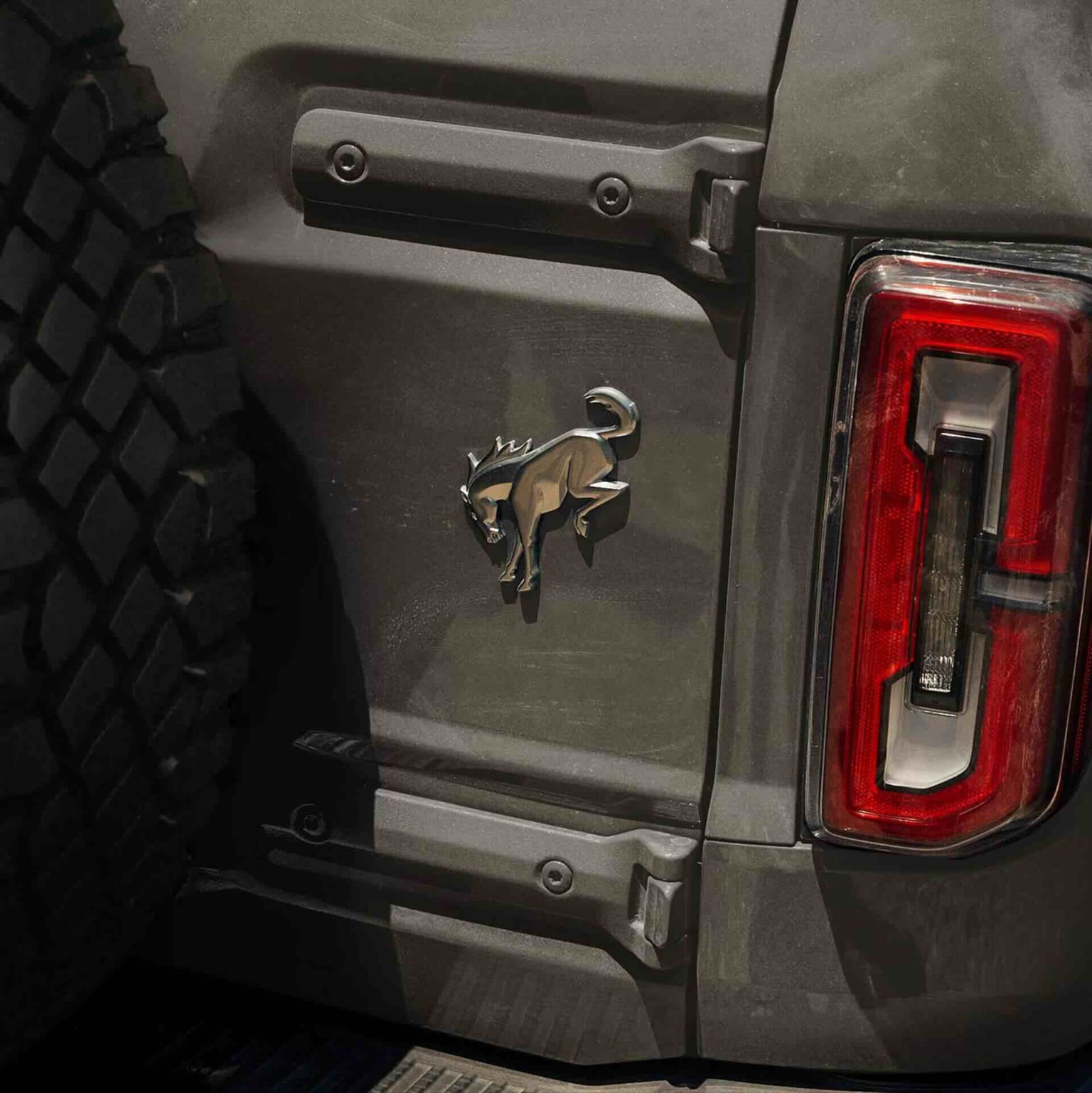 Close-up of a dark gray SUV's rear, showcasing a spare tire on the left and a taillight on the right, adorned with a chrome horse emblem in the center of the tailgate.