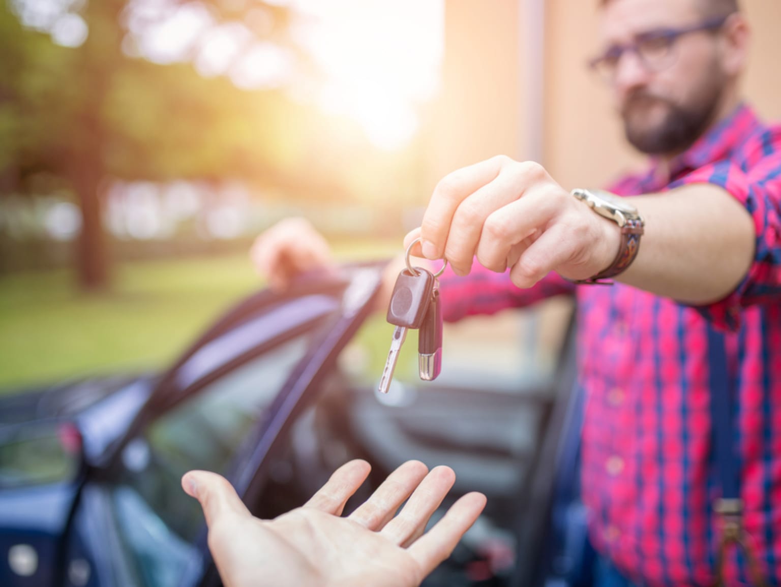 A person's hand holding car keys in the foreground, with a blurred vehicle in the background and a natural, outdoor setting.