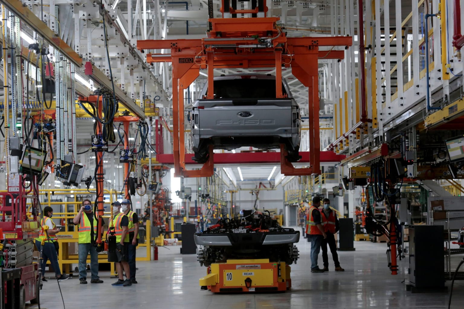 a car being lifted by a crane in a factory with workers standing in the foreground and a car being lifted by a crane in the background