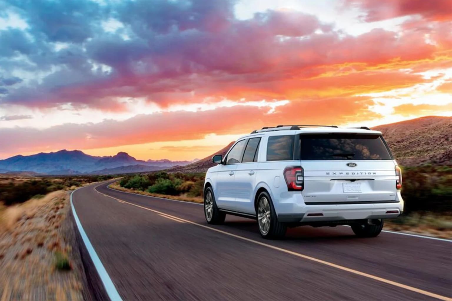A white SUV driving down a desert road with a dramatic, colorful sunset sky in the background.