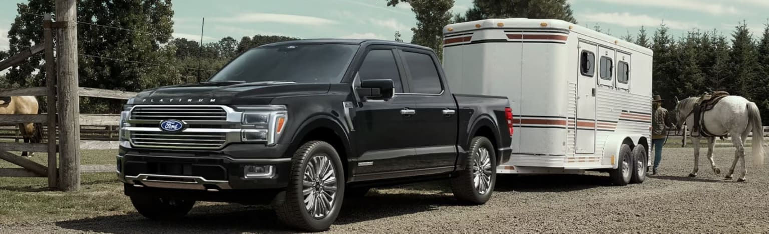 A black truck tows a white horse trailer with a horse and rider nearby, set against a backdrop of trees and a wooden fence.