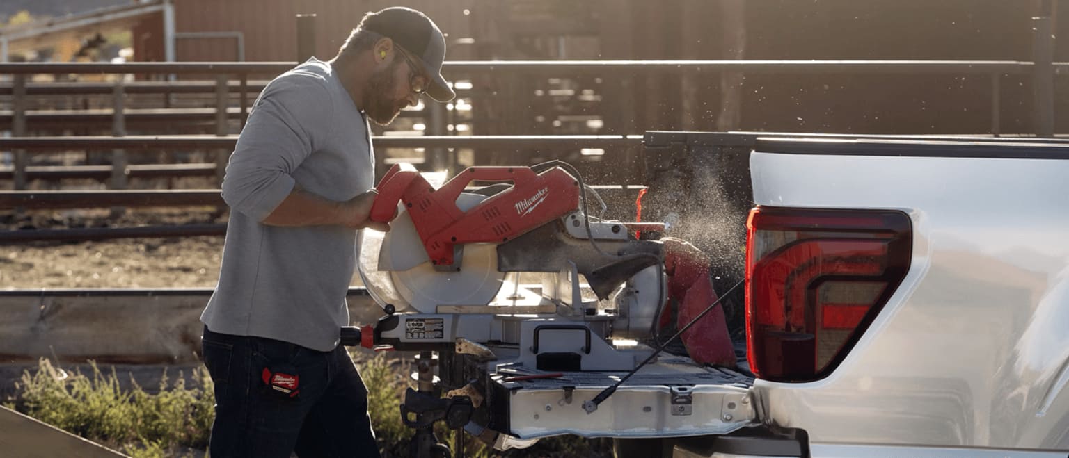 A mechanic in a gray shirt is working on a piece of machinery in a workshop setting, with various tools and equipment visible in the background.