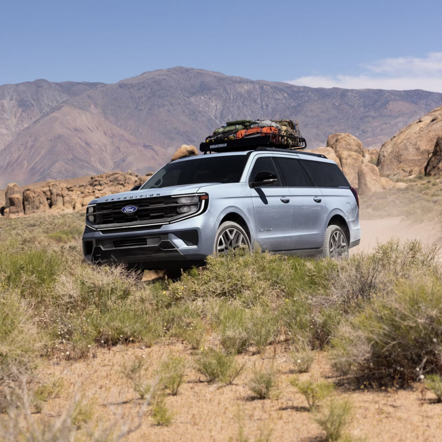 A light blue SUV with a roof rack loaded with gear drives on a dusty dirt road through a desert landscape with rocky hills and sparse vegetation under a clear blue sky.