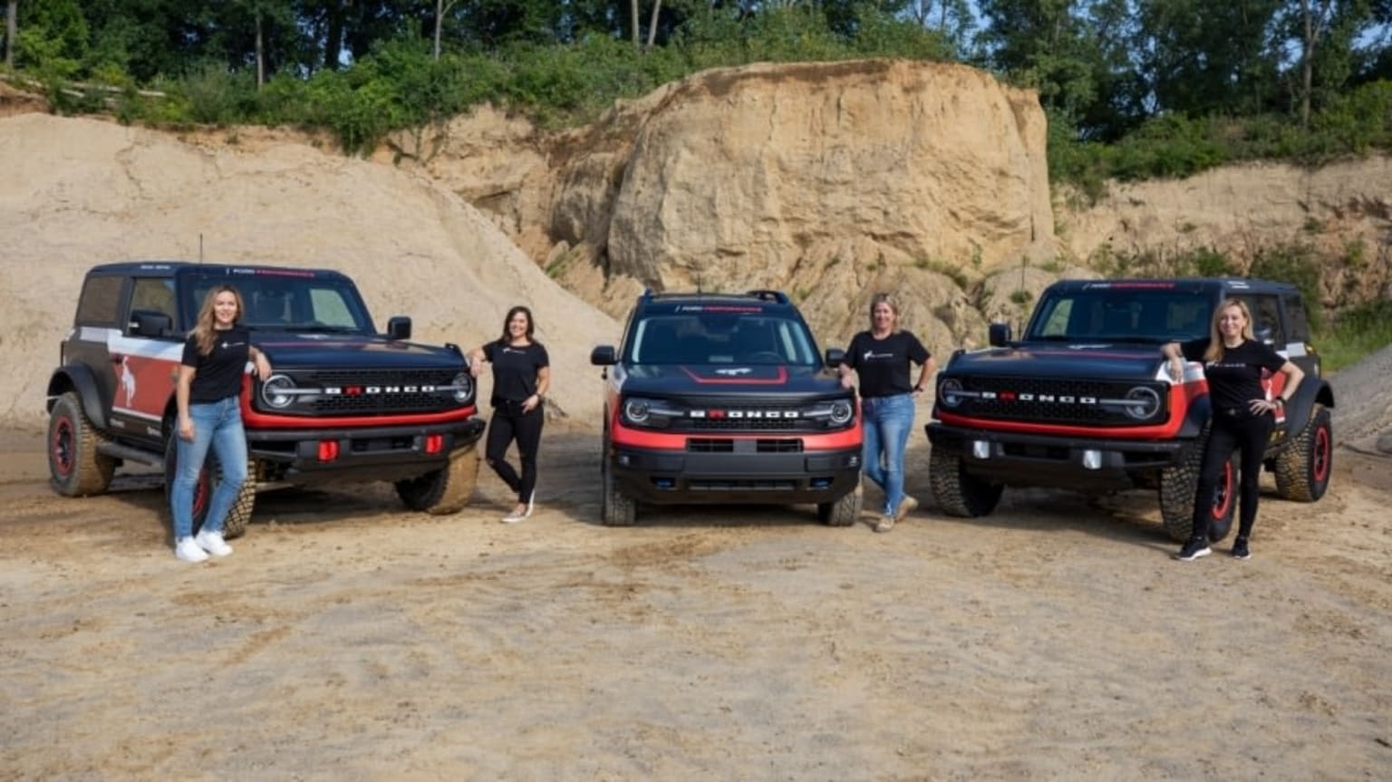 a group of people standing in front of four pickup trucks on a dirt road near a rock formation and trees