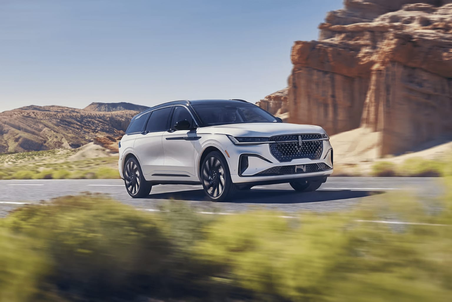 A sleek, white SUV driving on a winding road through rugged, mountainous landscape with rocky cliffs and lush vegetation in the background.