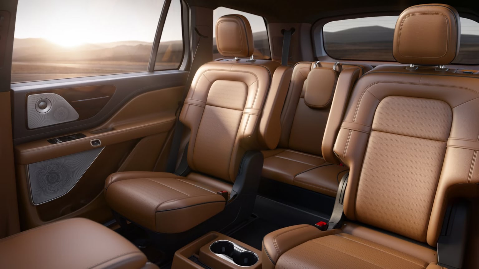 The image shows the interior of a luxury vehicle, with tan leather seats and a spacious cabin. The background appears to be a scenic landscape, with mountains and a warm, golden sunset visible through the windows.