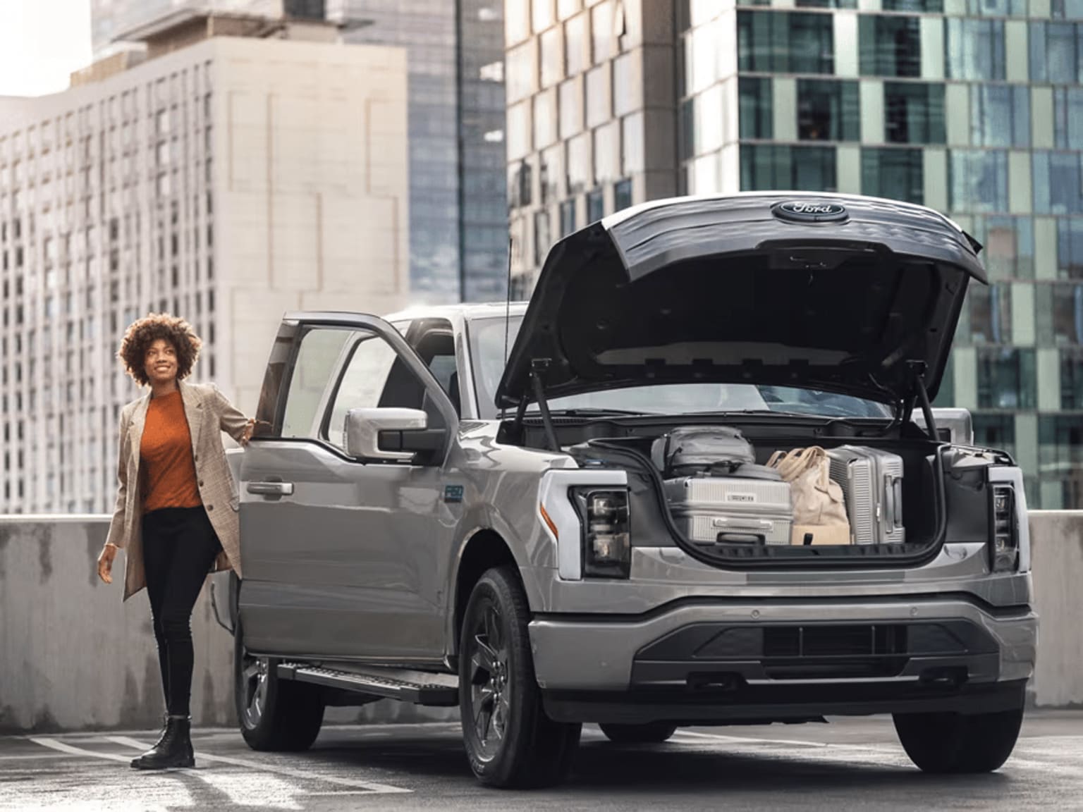 A woman standing next to a large silver pickup truck with the hood open in an urban setting.
