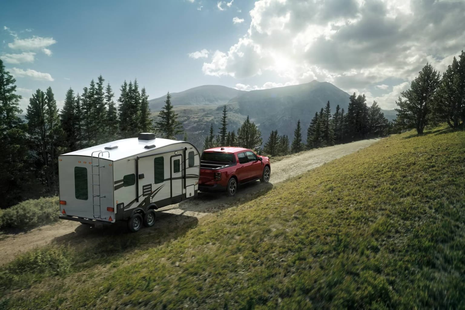 A red pickup truck towing a white travel trailer on a dirt road through a pine forest, with a mountain range in the background under a cloudy sky.