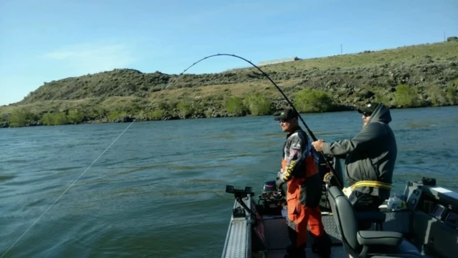 Two fishermen in a boat reel in a catch on a wide river with a rocky, vegetated hillside in the background under a clear blue sky.