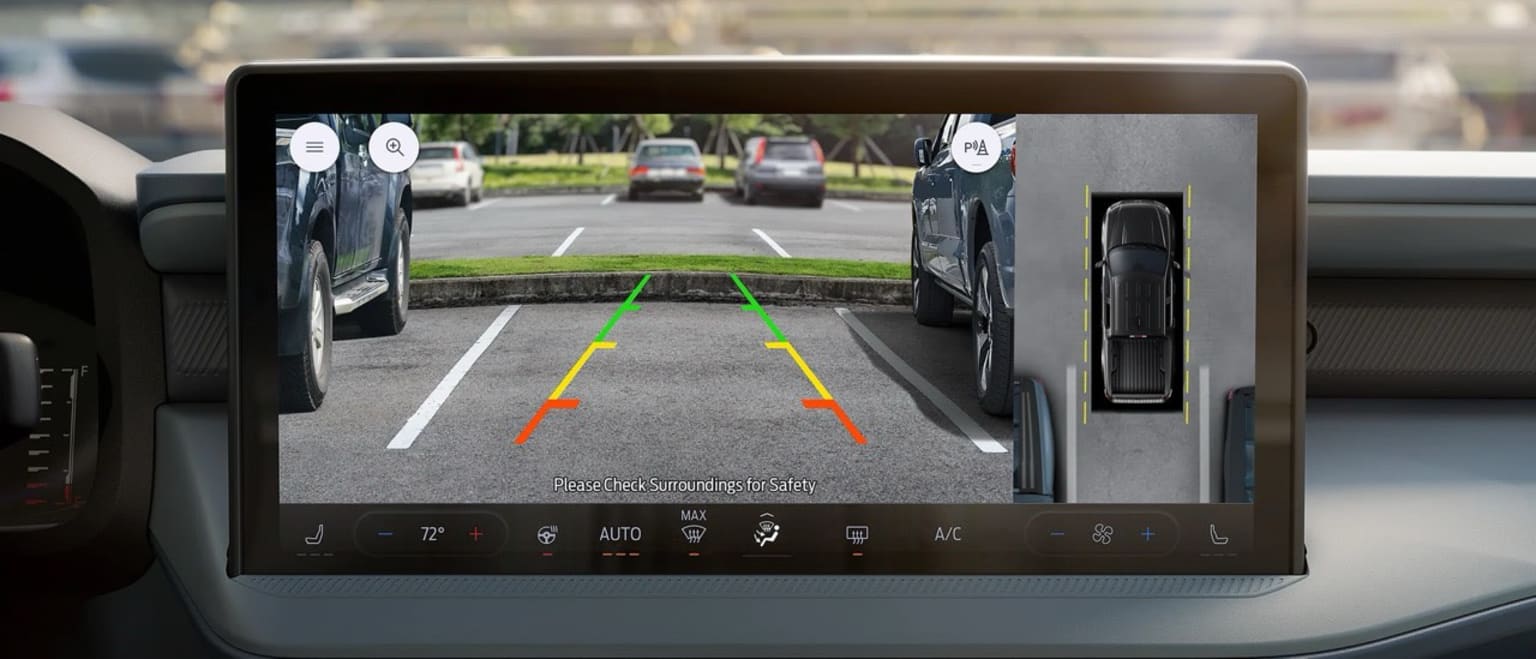 The image depicts a dashboard display showing a rear-view camera view of the road ahead. Various indicators and controls are displayed, with a paved street and vehicles visible in the background.