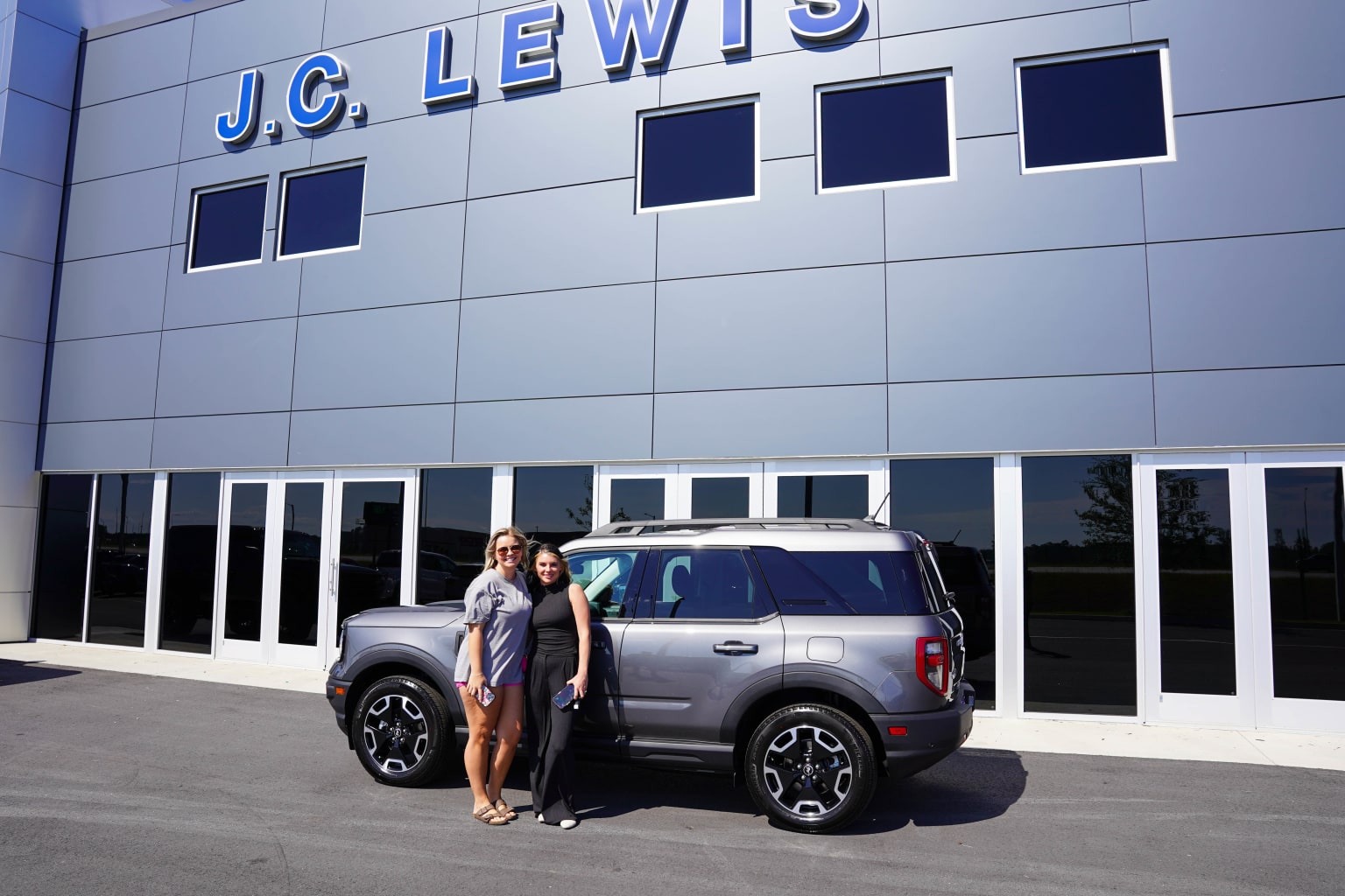 Two women standing in front of a new Ford Bronco with the J.C. Lewis Ford Pooler dealership in the background.