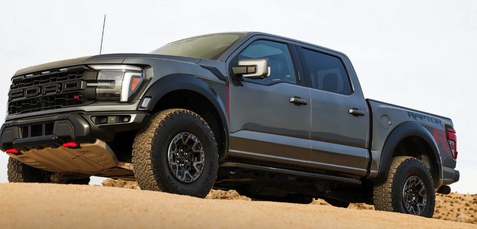 A large, rugged-looking pickup truck with off-road tires and a bold, aggressive design sits on a sandy, desert-like terrain in the foreground.