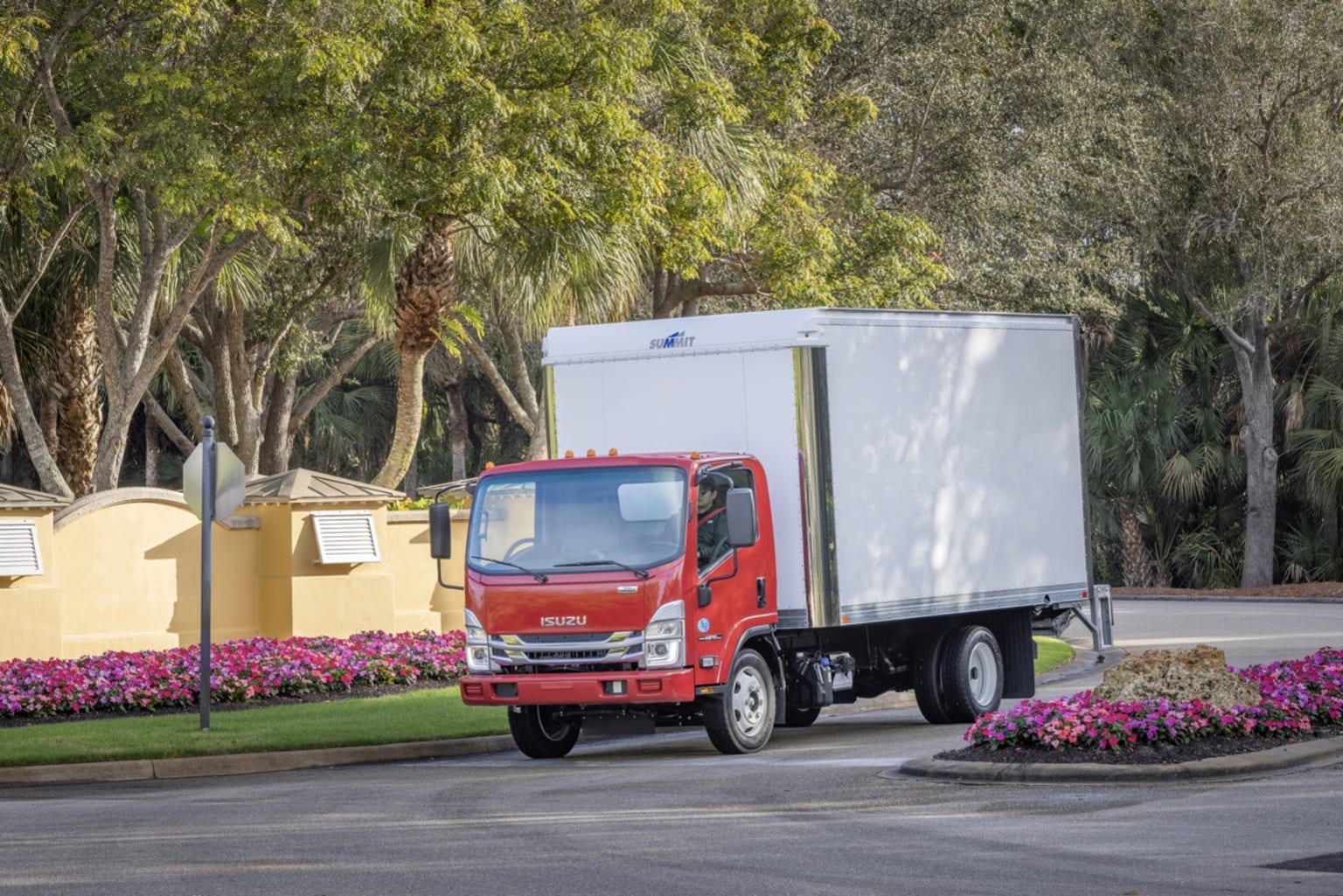 A red box truck drives on a paved road with manicured bushes and trees in the background.