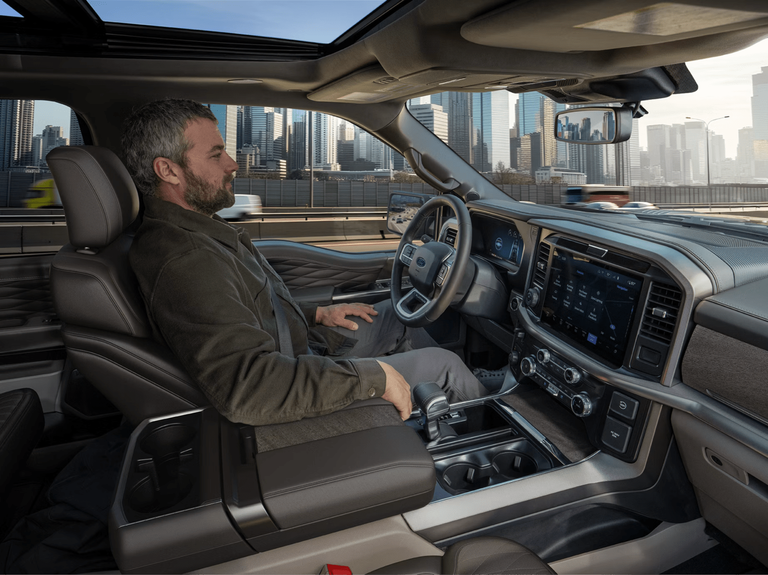 A person sits in the driver's seat of a modern vehicle interior with a city skyline visible through the windows.