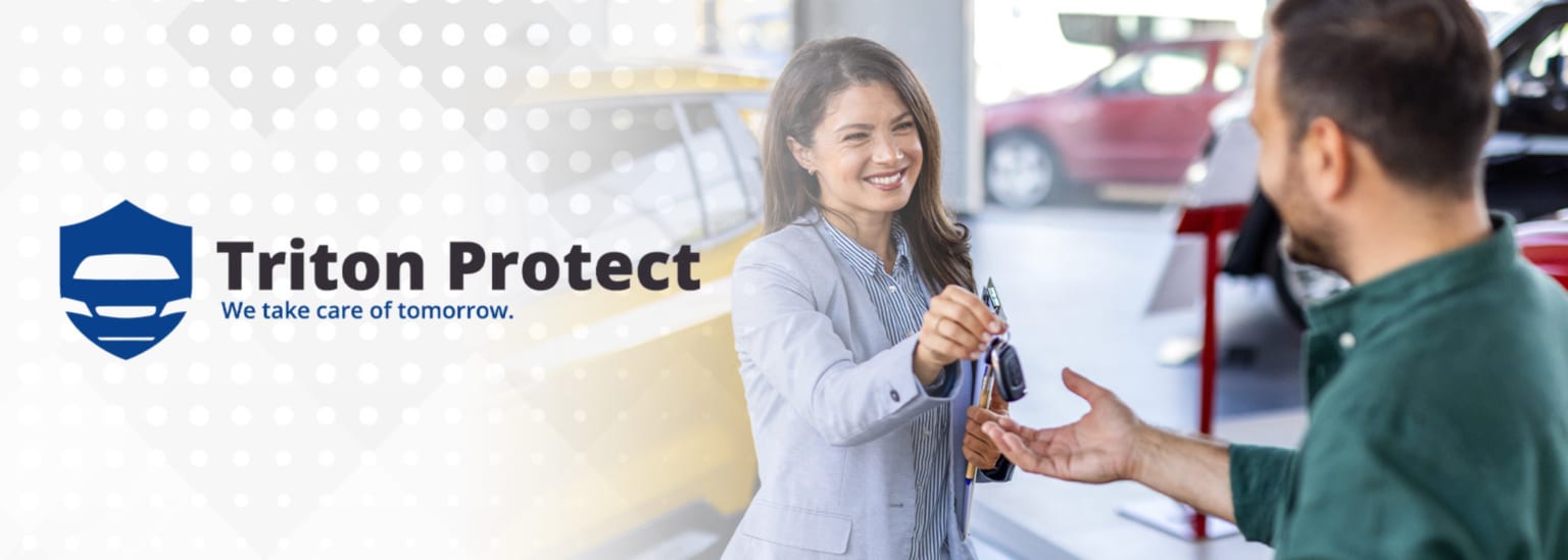 A smiling woman hands car keys to a man in the foreground, with various cars visible in the blurred background of a dealership.