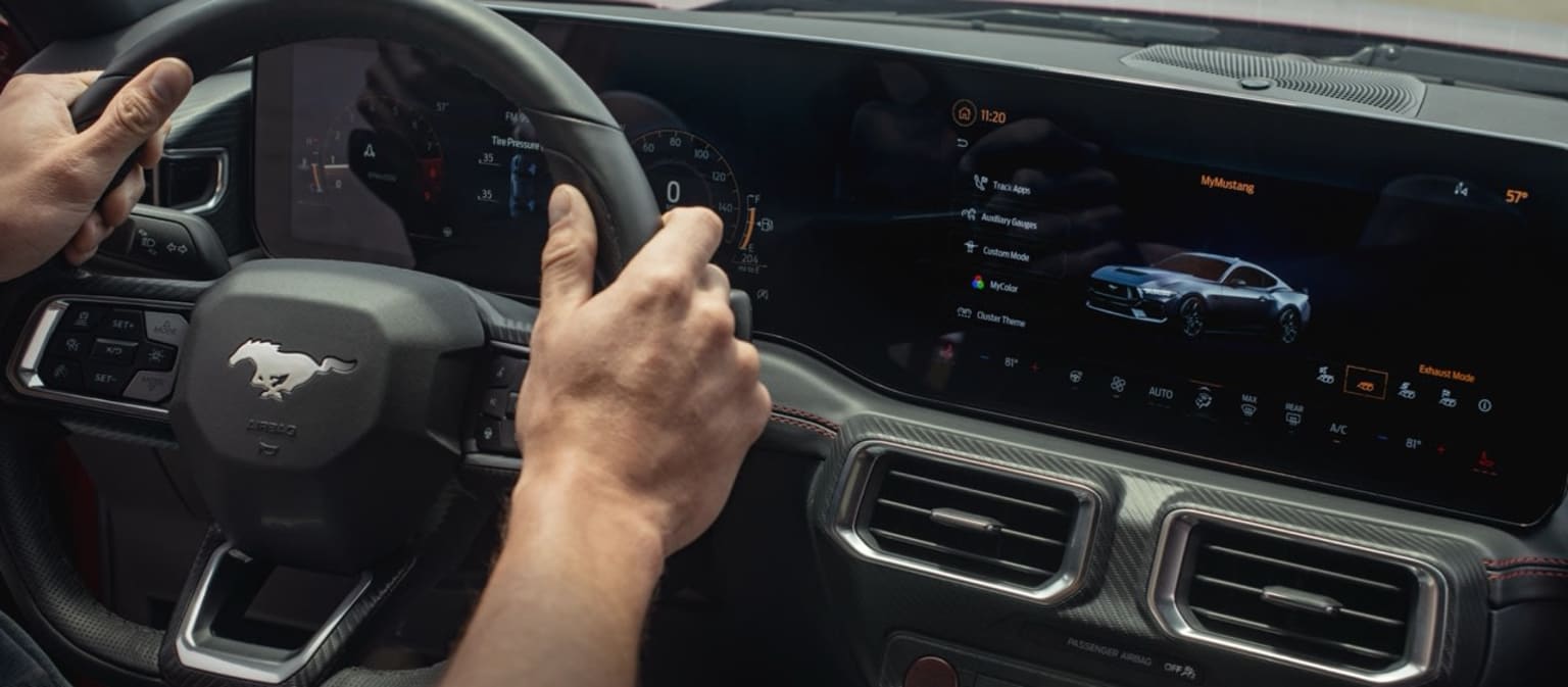 The image depicts a car interior with a person's hands on the steering wheel and a digital display in the background displaying various vehicle information and controls.