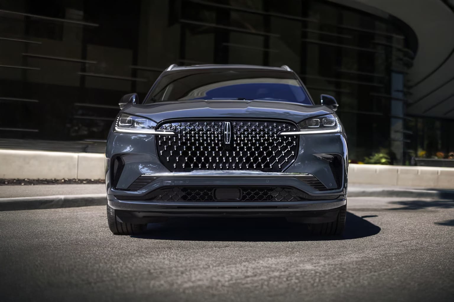 A sleek, dark-colored SUV with a prominent grille and headlights stands in the foreground, against a backdrop of a building and a paved surface.