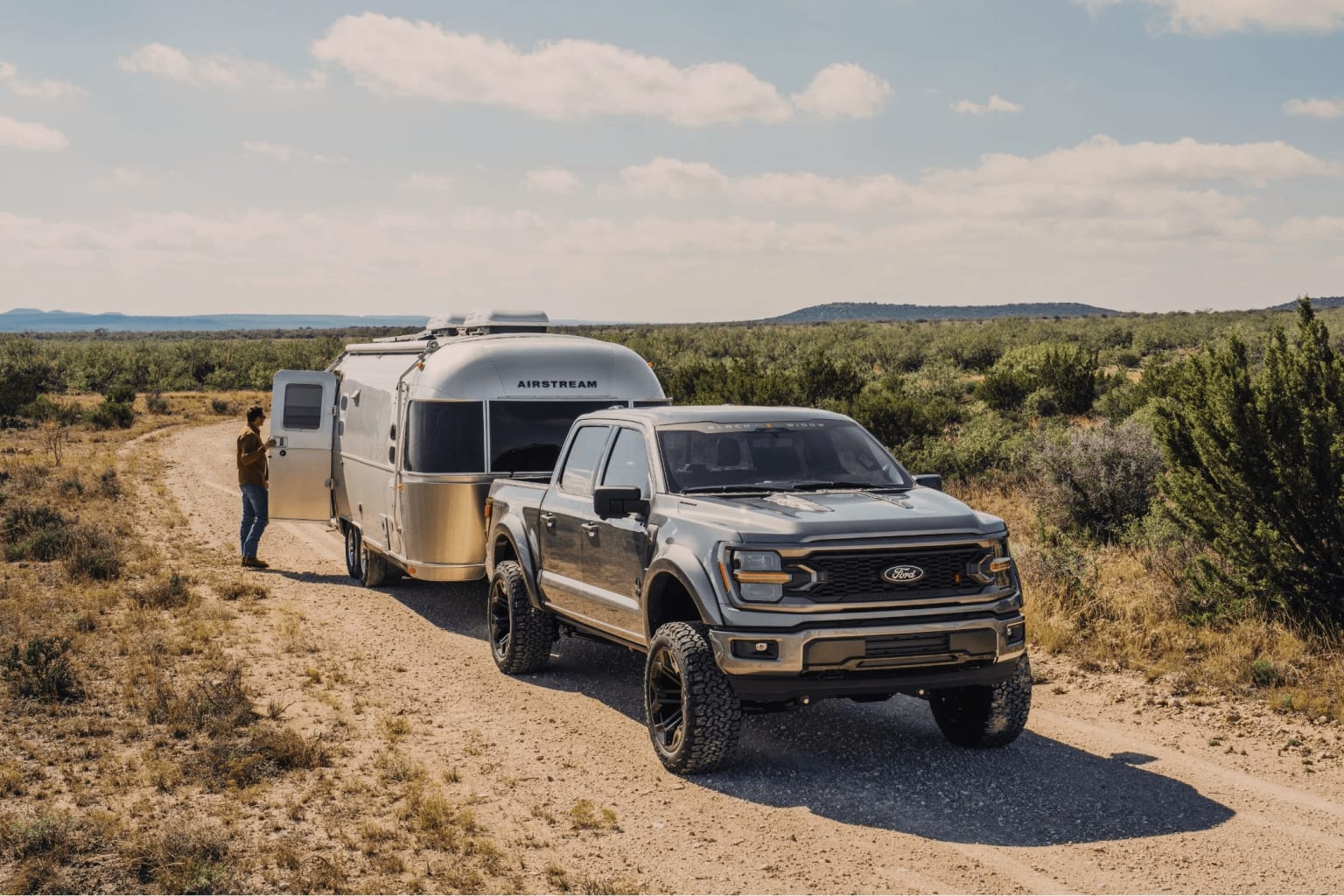 A pickup truck towing a camper trailer on a dirt road in a desert landscape with hills and mountains in the background.