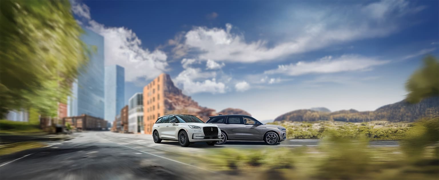 A white luxury SUV parked on a city street with modern buildings and a mountainous background.