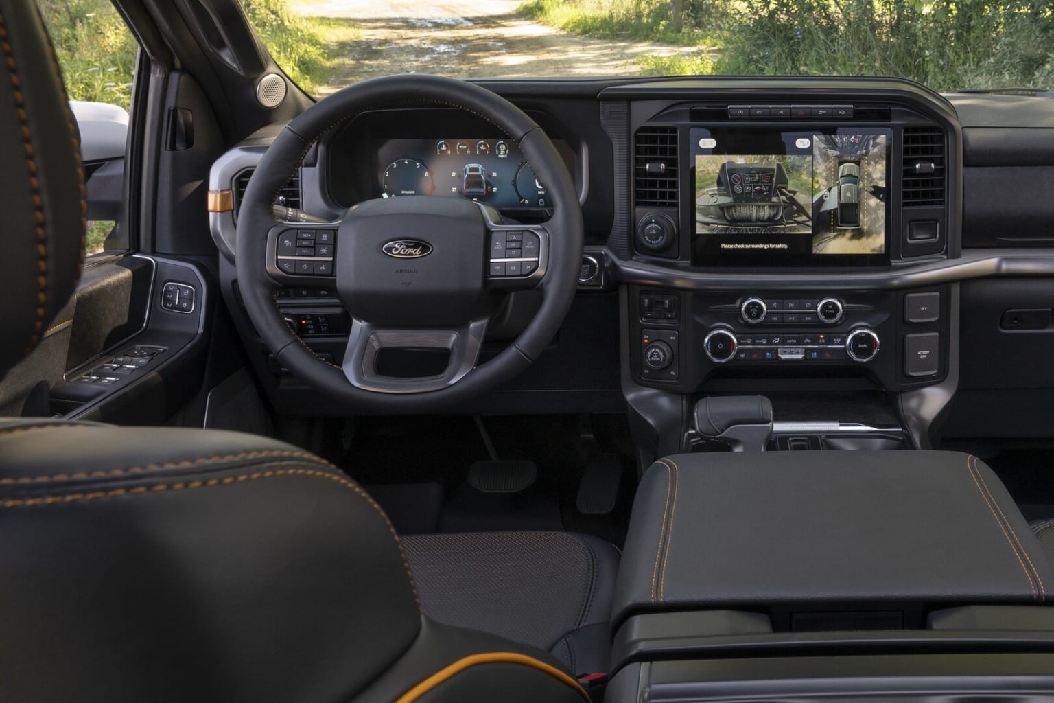 Interior view of a vehicle with dashboard, steering wheel, and various controls and displays. Background showcases rural or natural setting with trees and greenery visible through the windshield.