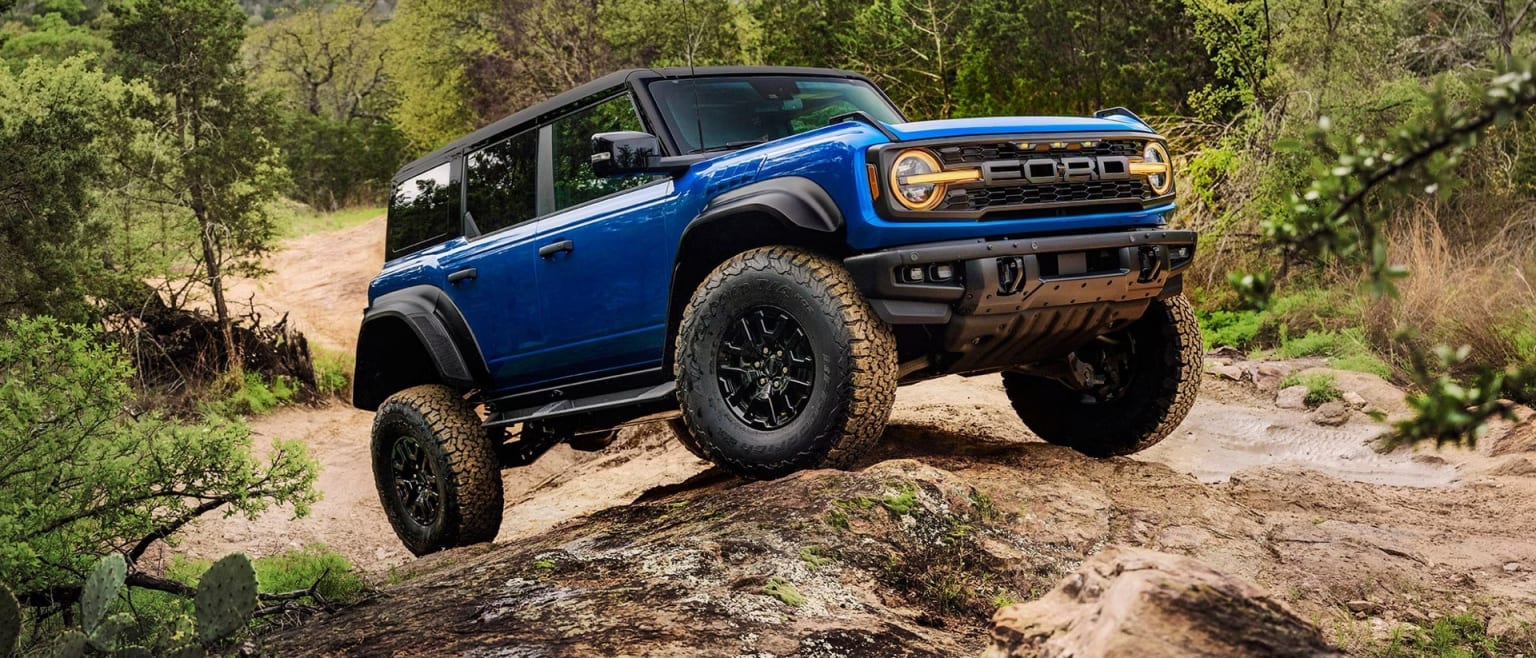 A blue SUV is navigating a rocky, off-road trail surrounded by lush green trees and vegetation.
