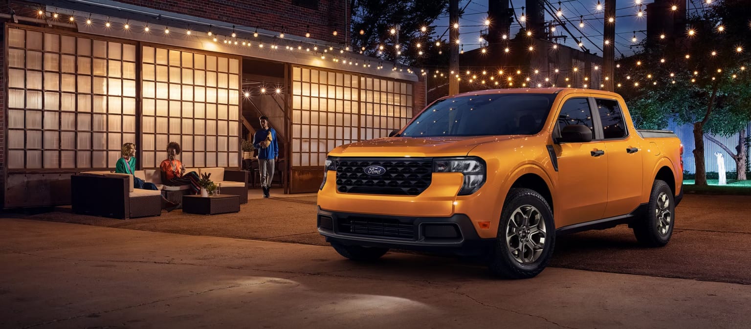 A yellow pickup truck parked in an outdoor setting surrounded by string lights and a brick building.