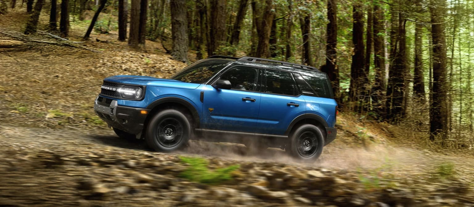 A blue SUV driving on a dirt path through a forest with tall trees.
