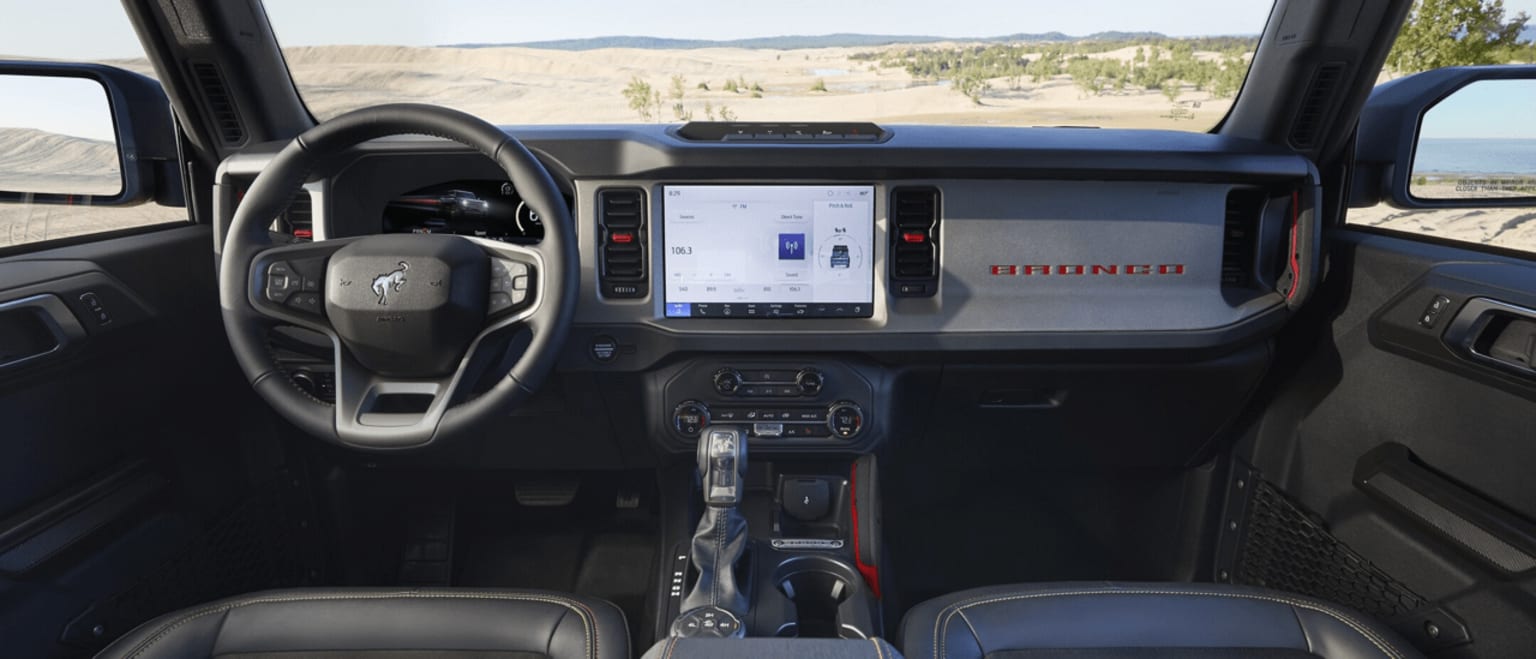 The image depicts the interior of a vehicle with a steering wheel, dashboard, and a large touchscreen display in the center console. The background outside the vehicle shows a desert landscape with mountains in the distance.