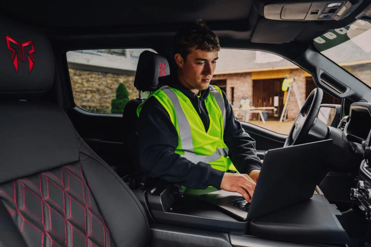A person in a high-visibility vest using a laptop in the driver's seat of a vehicle.