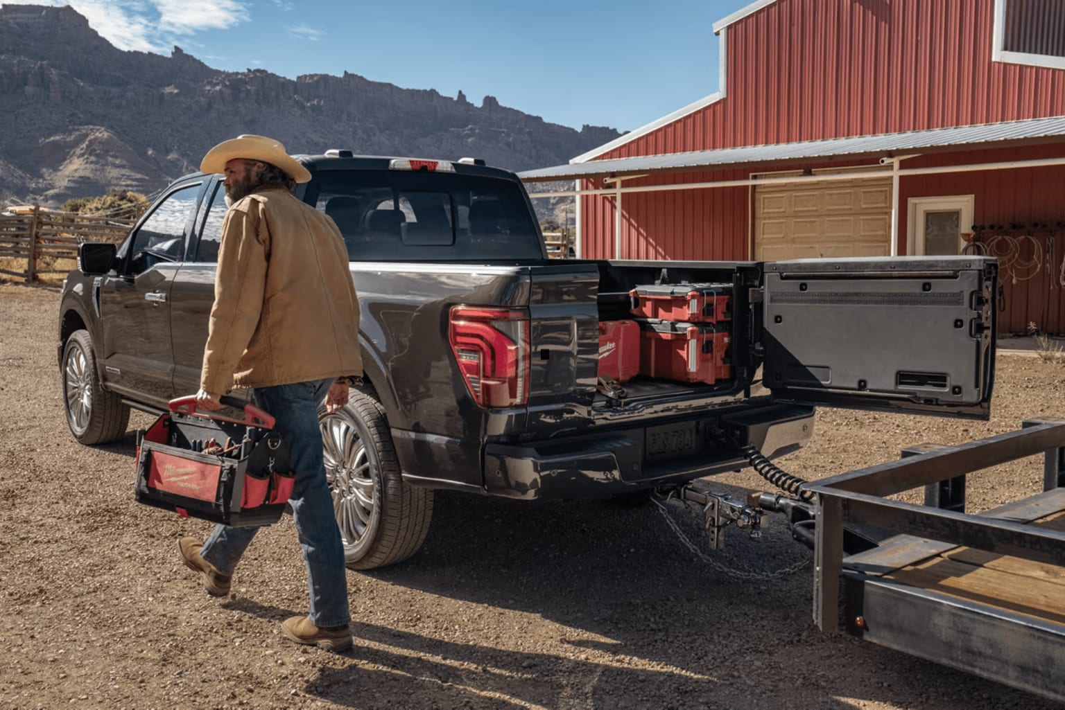 A rugged pickup truck with various outdoor gear and equipment parked in front of a red barn-like structure, set against a backdrop of mountainous terrain.