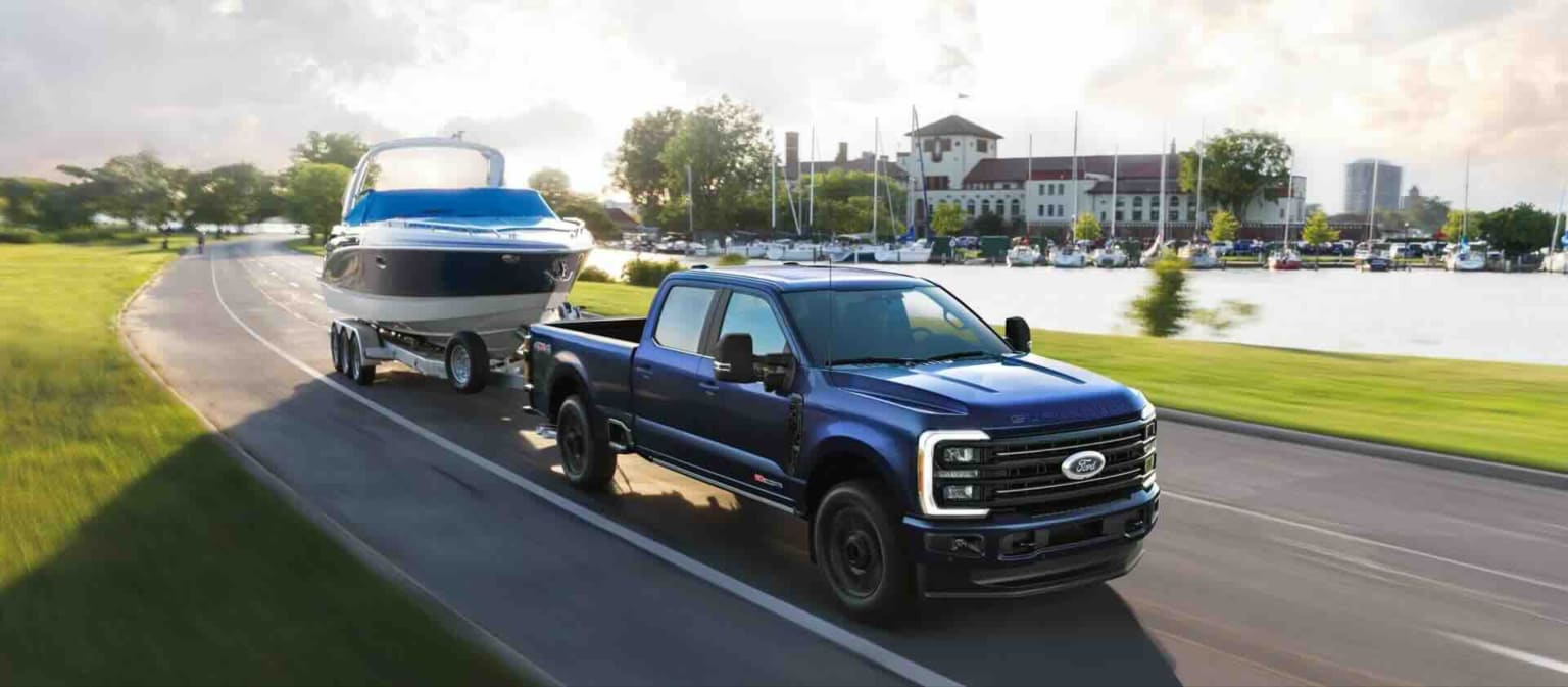 A blue truck towing a boat on a trailer with a waterfront town and marina in the background.