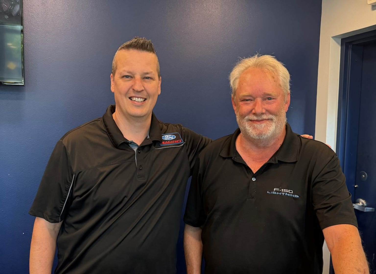 Two men standing in front of a blue wall, one younger with short blond hair and the other older with a long gray beard, both wearing black polo shirts.