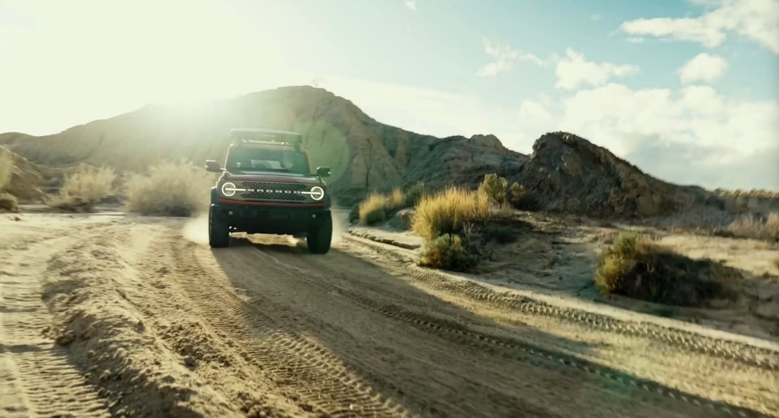 Off-road vehicle driving on a dusty mountain road with rocky peaks and a bright sky.