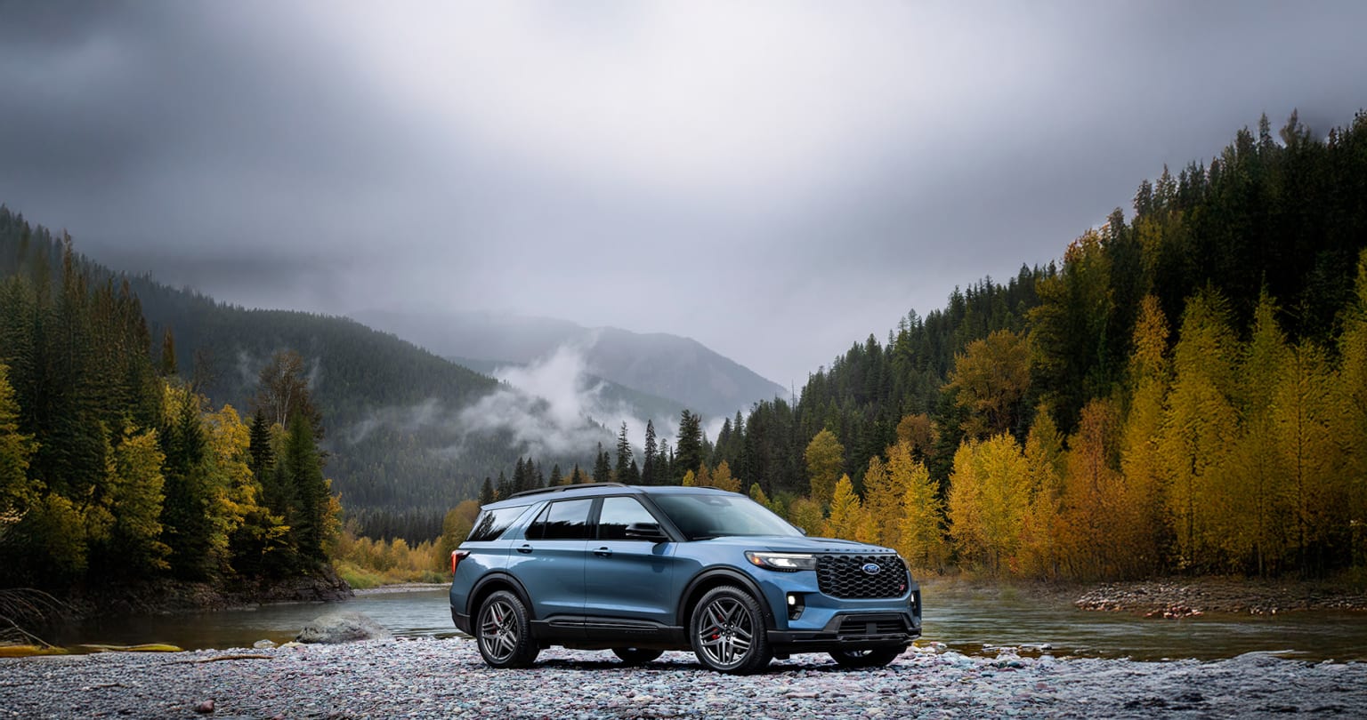 SUV in nature scene with rocky riverbed, autumnal forest, and fog-shrouded mountains.