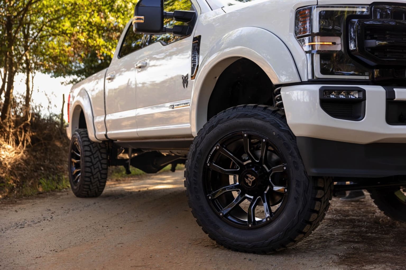 A large, lifted white pickup truck with large off-road tires and black rims on a dirt path surrounded by lush, green foliage.