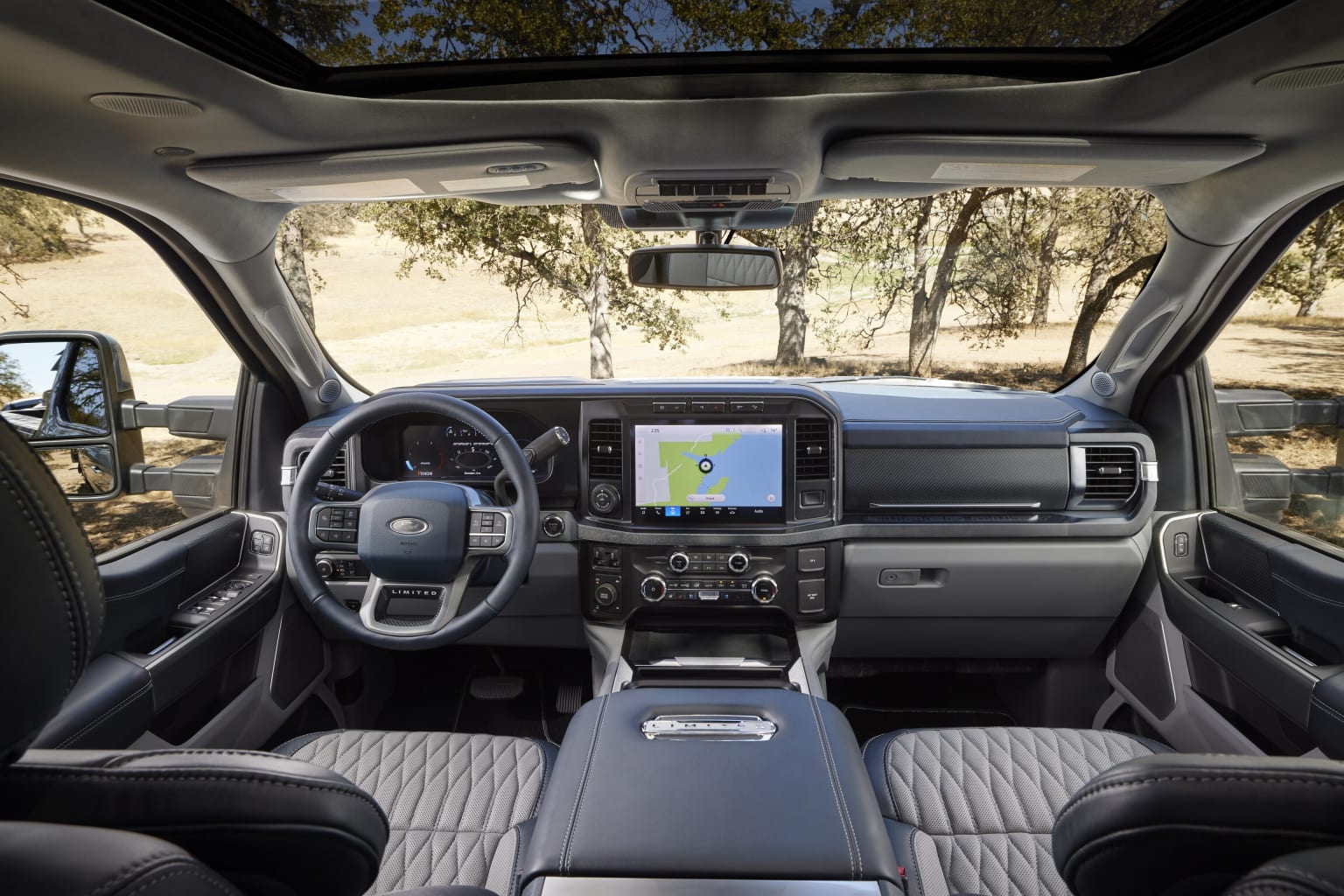 Interior of a modern vehicle with a dashboard displaying a touchscreen, steering wheel, and controls. Background shows a wooded area with trees.