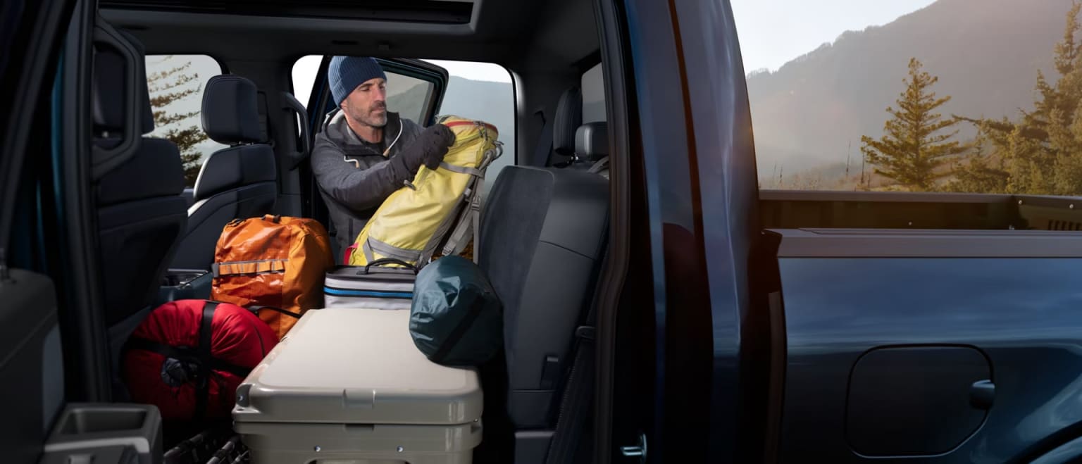 A man loads camping gear into the back of a truck with a mountainous, forested landscape in the background.