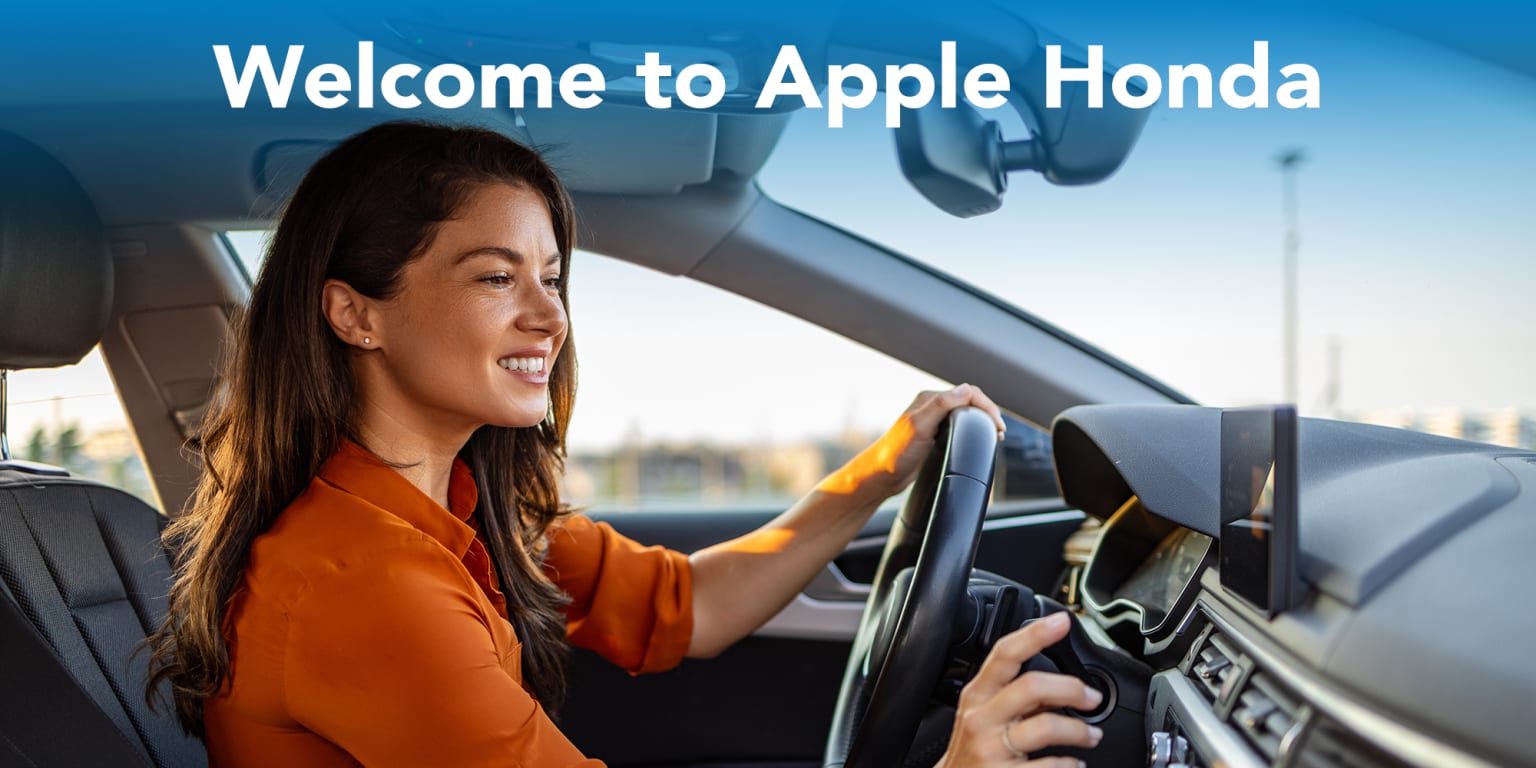 A woman in an orange shirt driving a car with a bright sky in the background.