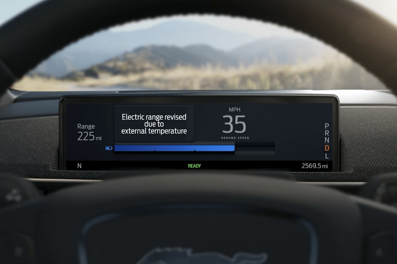 The image shows the dashboard of a vehicle with displays of current speed, range, and external temperature, with a mountainous landscape visible through the windshield.