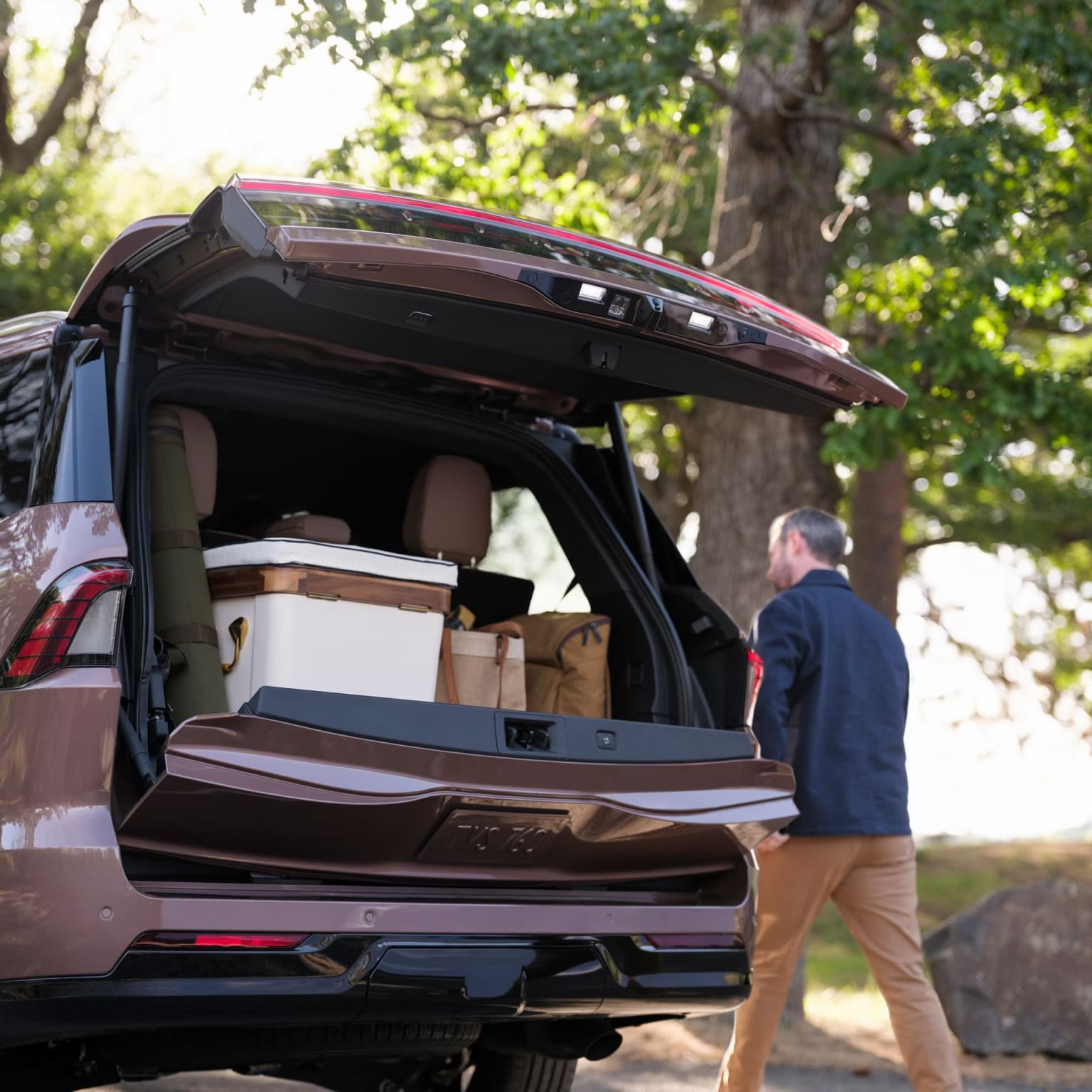 A burgundy SUV with its rear hatch open, revealing a cargo area filled with boxes, stands in a wooded area with trees and sunlight filtering through the foliage in the background.