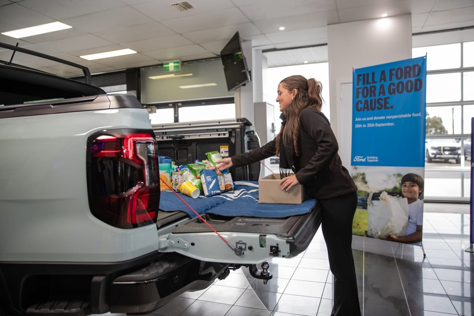 A woman is loading items into the back of a vehicle in a car dealership showroom, with a large banner promoting a charity campaign visible in the background.