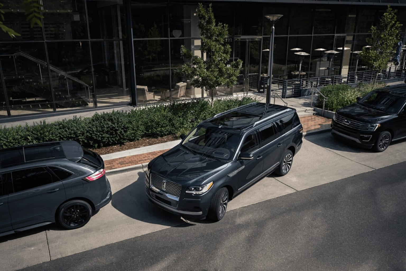 The image is of a luxury SUV parked on a sidewalk outside a modern building with glass walls and lush greenery.